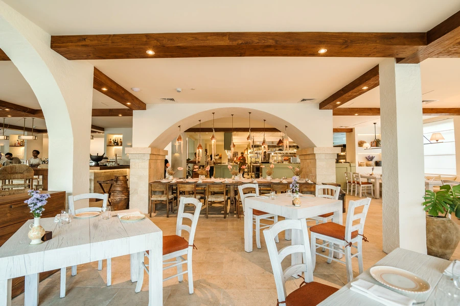 Interior of Zia Tina Restaurant featuring white arched doorways, rustic wooden ceiling beams, and a mix of long wooden communal tables and smaller white dining sets